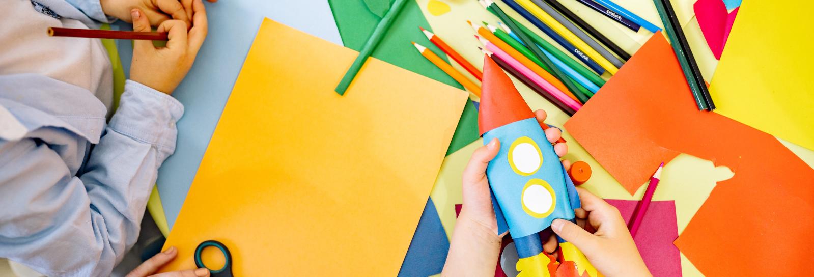 Børns hænder rundt om et børn med karton og farveblyanter. Nederst til højre er en person i gang med a lave en raket ud af karton.