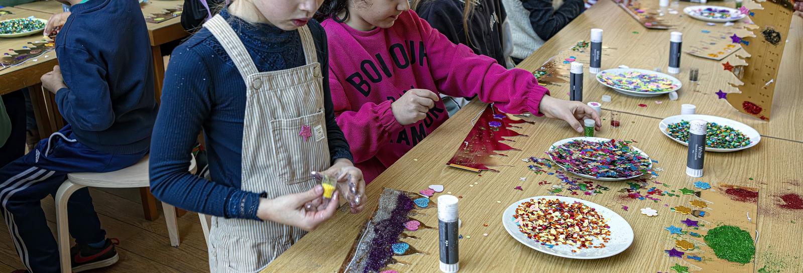 To børn står ved et bord, og er i gang med at pynte en udstanset pap-krone der ligger foran dem. På bordet omkring dem er der tallerkener med pailletter, glimmer og limstifter.