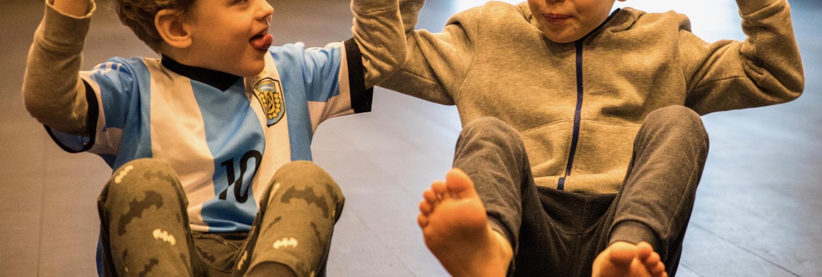 To store børnehavebørn laver siddende balanceøvelse på gulvet